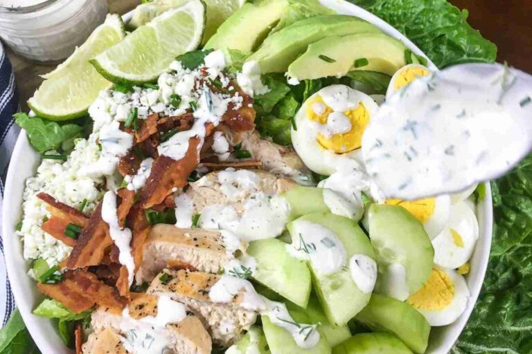 A bowl of salad with grilled chicken, bacon, hard-boiled eggs, avocado, cucumber, lettuce, cheese, lime wedges, and ranch dressing being spooned on top.