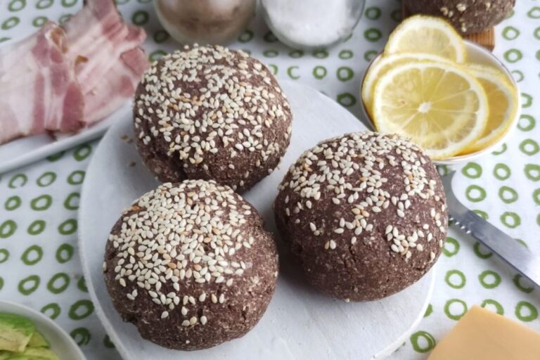 Three sesame-seeded brown bread rolls on a white plate, surrounded by lemon slices, bacon, and a knife with a polka dot tablecloth underneath.