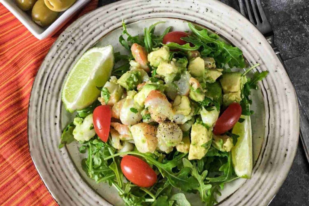 A plate of salad with shrimp, avocado, arugula, cherry tomatoes, and lime wedges, served on a ceramic dish with a fork and a bowl of green olives in the background.