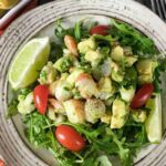A plate of salad with shrimp, avocado, arugula, cherry tomatoes, and lime wedges, served on a ceramic dish with a fork and a bowl of green olives in the background.