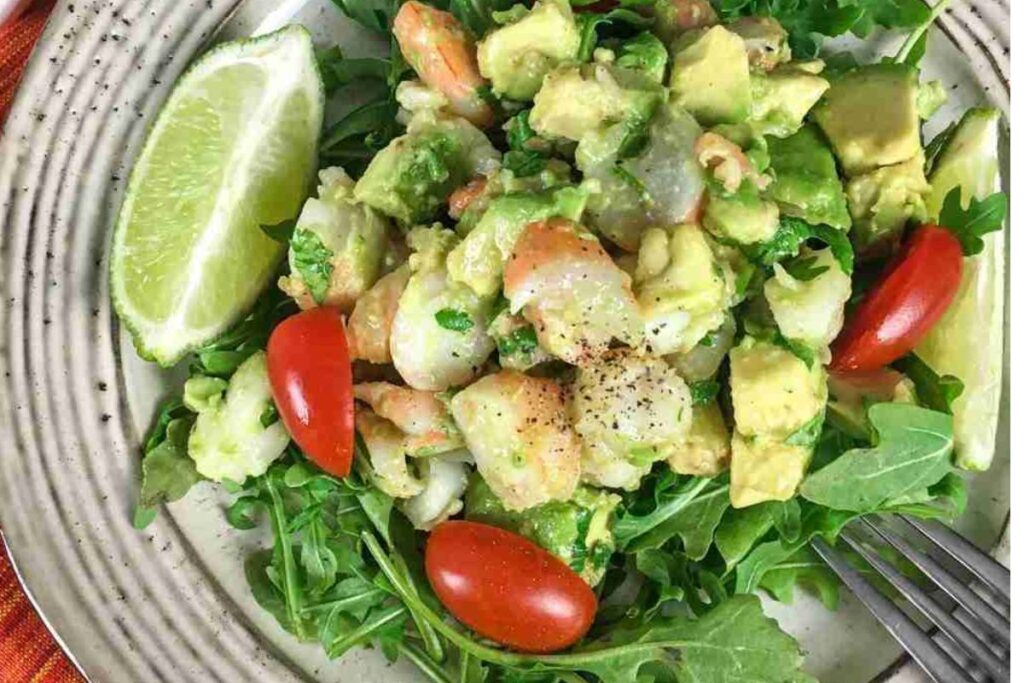 A plate of salad with shrimp, avocado, cherry tomatoes, arugula, and a lime wedge, seasoned with black pepper.