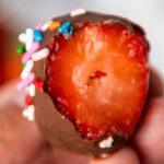 A bitten chocolate-covered strawberry is held between fingers. The strawberry has colorful sprinkles on the chocolate coating.