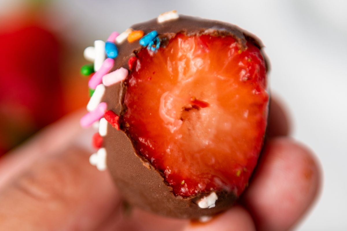 A bitten chocolate-covered strawberry is held between fingers. The strawberry has colorful sprinkles on the chocolate coating.