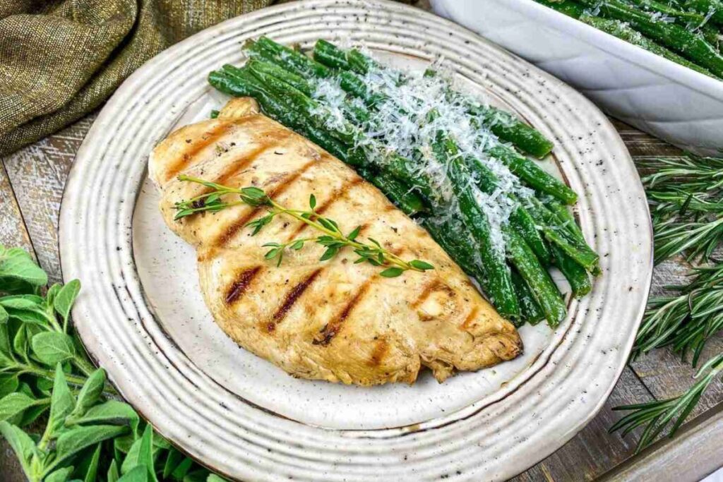 Grilled chicken breast garnished with herbs served alongside green beans topped with grated cheese on a white plate.