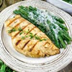 Grilled chicken breast garnished with herbs served alongside green beans topped with grated cheese on a white plate.