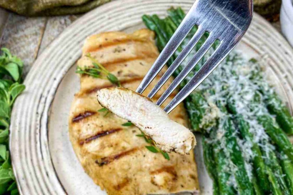 A fork holds a slice of grilled chicken breast above a plate with green beans topped with grated cheese.