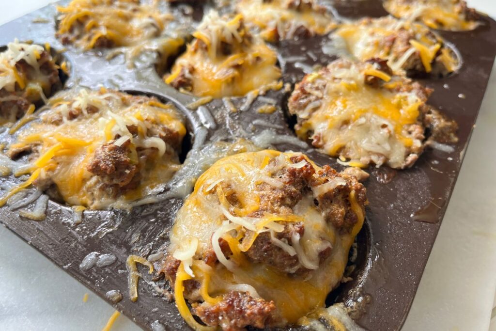 A close-up of a muffin tin filled with baked cheesy ground beef cups topped with melted shredded cheese.