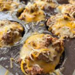 A close-up of a muffin tin filled with baked cheesy ground beef cups topped with melted shredded cheese.
