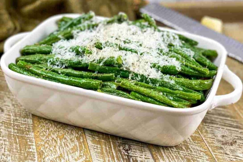 A white baking dish filled with cooked green beans topped with grated cheese, placed on a wooden surface.