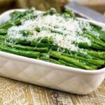 A white baking dish filled with cooked green beans topped with grated cheese, placed on a wooden surface.