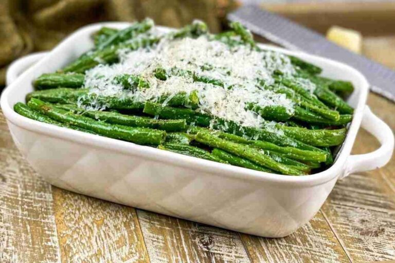 A white baking dish filled with cooked green beans topped with grated cheese, placed on a wooden surface.