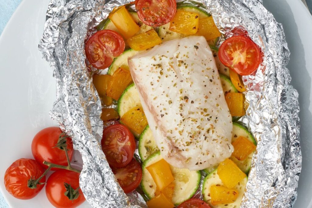 A piece of white fish seasoned with herbs, baked on zucchini, cherry tomatoes, and yellow bell peppers, served in an open foil packet with extra tomatoes on the side.
