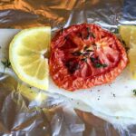 A raw white fish fillet on aluminum foil topped with a slice of tomato, two lemon wedges, black pepper, and fresh thyme.