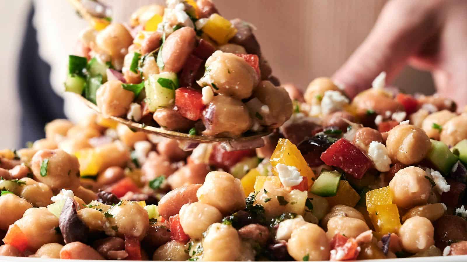 A spoonful of chickpea salad with chopped cucumbers, tomatoes, peppers, parsley, and feta cheese being served from a bowl.