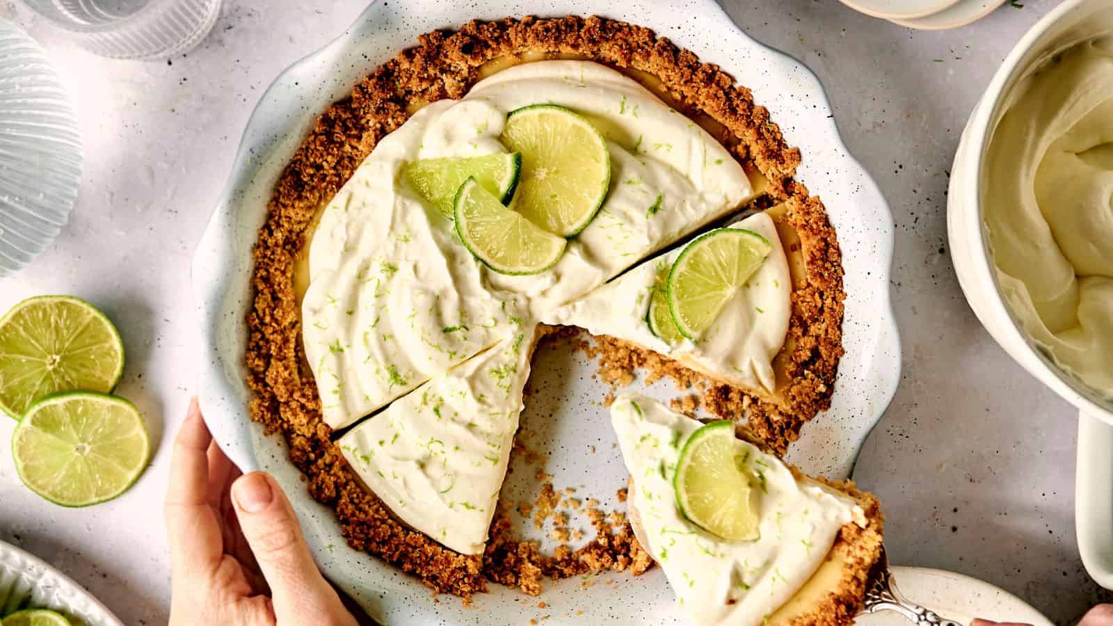 Key lime pie with a slice removed on a white plate. Topped with lime wedge and zest, the crust is golden brown. A hand touches the plate edge, and a small bowl with cream is nearby.