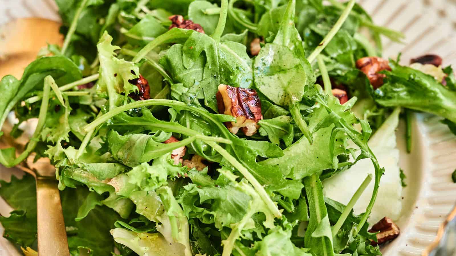 A close-up of a fresh green salad with mixed leafy greens, cucumber slices, and pecans on a plate with a gold fork.