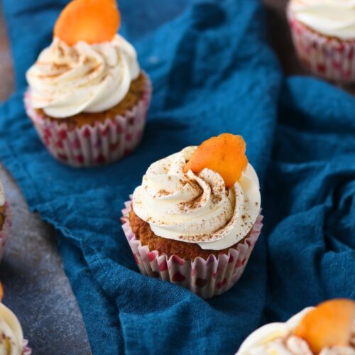 Keto carrot cake cupcakes topped with cream cheese frosting and a small carrot garnish on a blue cloth.