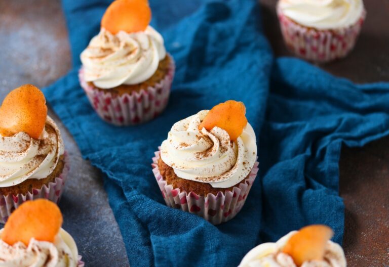 Keto carrot cake cupcakes topped with cream cheese frosting and a small carrot garnish on a blue cloth.