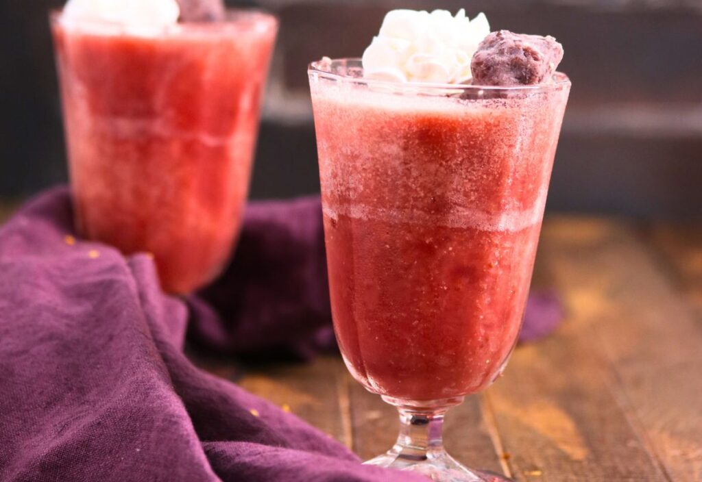 A glass of strawberry jello drink topped with whipped cream and a cherry, set on a wooden surface with a purple cloth nearby. Another similar drink is in the background.