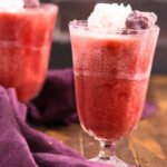 A glass of strawberry jello drink topped with whipped cream and a cherry, set on a wooden surface with a purple cloth nearby. Another similar drink is in the background.