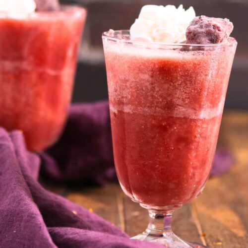 A glass of strawberry jello drink topped with whipped cream and a cherry, set on a wooden surface with a purple cloth nearby. Another similar drink is in the background.