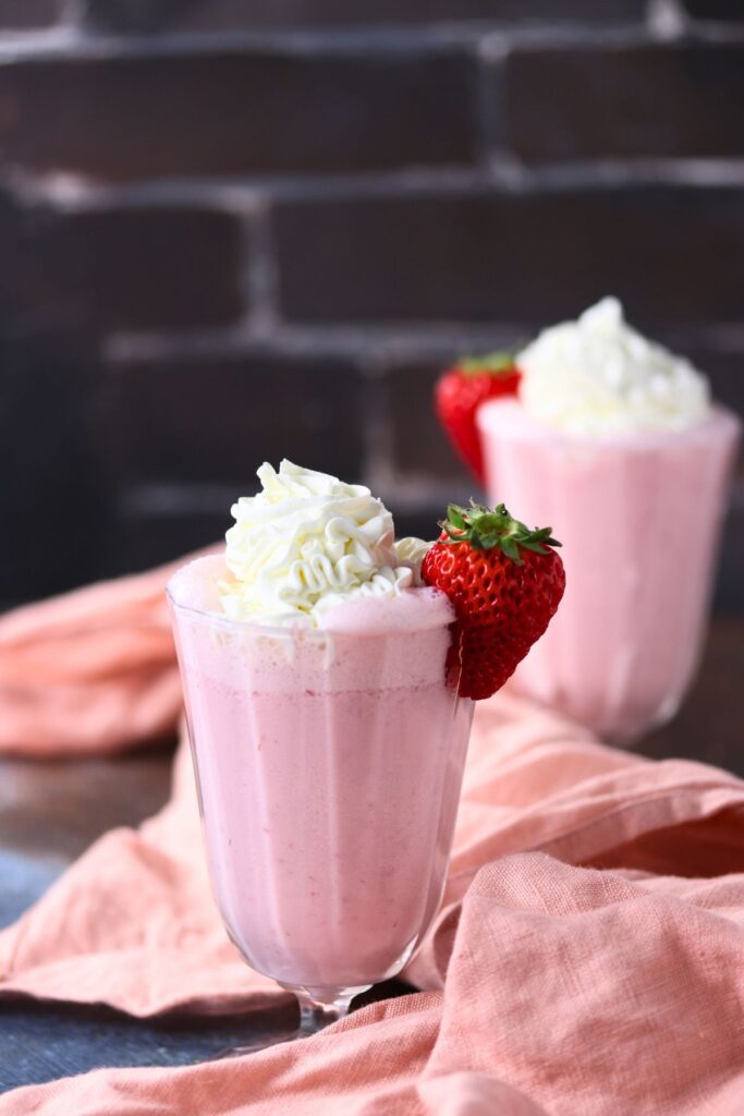 Two glasses of pink strawberry smoothie milkshake topped with whipped cream and garnished with a whole strawberry, set on a pink cloth with a dark brick background.