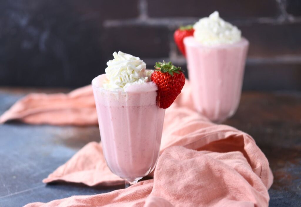 Two glasses of pink strawberry smoothie topped with whipped cream and a whole strawberry, placed on a table with a soft pink cloth.
