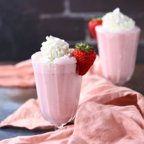 Two glasses of pink strawberry smoothie topped with whipped cream and a whole strawberry, placed on a table with a soft pink cloth.