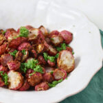 A white plate with roasted radish pieces garnished with chopped parsley, placed on a green cloth napkin.