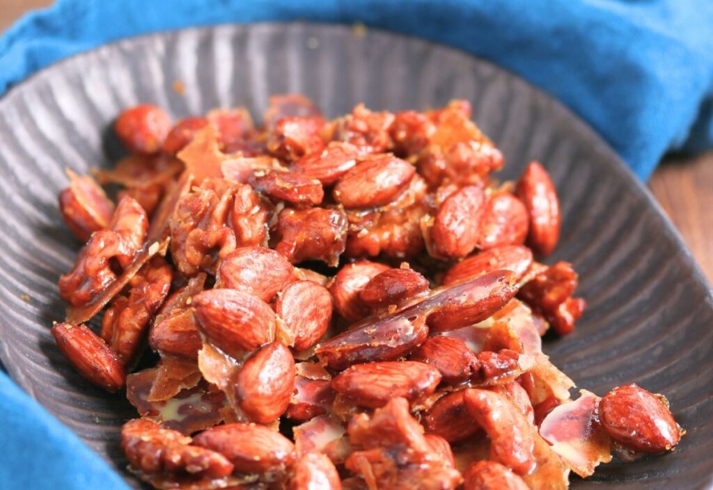 A black plate filled with clusters of caramelized nuts, including almonds and walnuts, sits on a blue cloth.