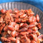 A black plate filled with clusters of caramelized nuts, including almonds and walnuts, sits on a blue cloth.