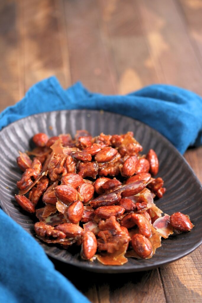 A black plate filled with glazed roasted almonds sits on a wooden surface with a blue cloth partially underneath.