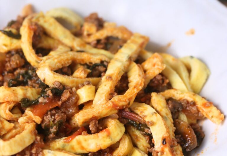 Close-up of pasta noodles mixed with ground meat, vegetables, and tomato sauce on a white plate.