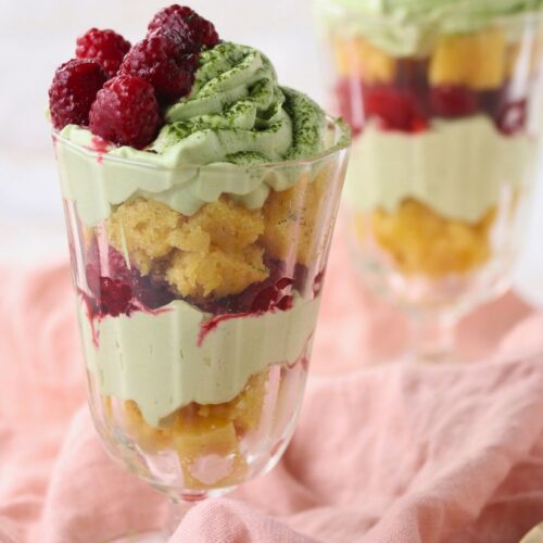 A glass dessert parfait with layers of green matcha cream, yellow sponge cake, raspberries, and berry sauce, placed on a pink cloth.