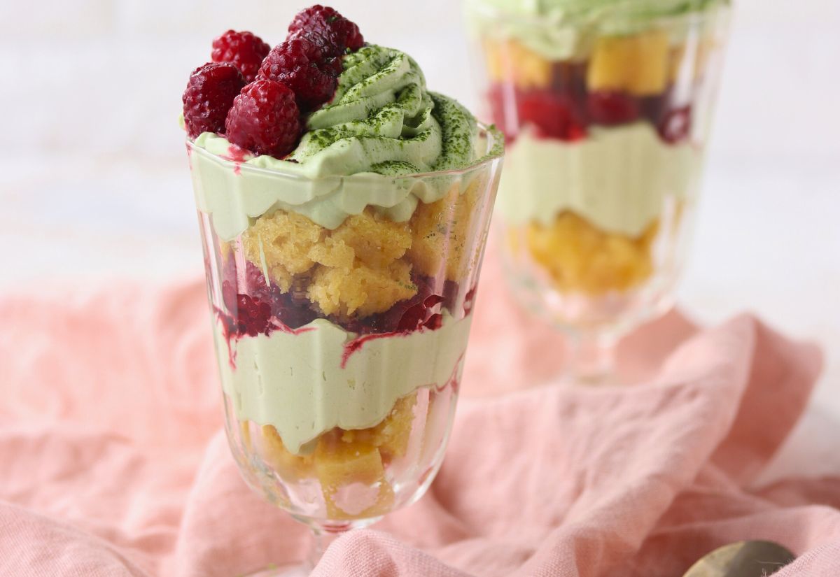 A glass dessert parfait with layers of green matcha cream, yellow sponge cake, raspberries, and berry sauce, placed on a pink cloth.