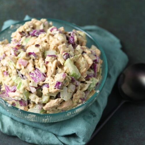 A glass bowl of chicken salad with chopped celery and red onion sits on a green cloth next to a black spoon.