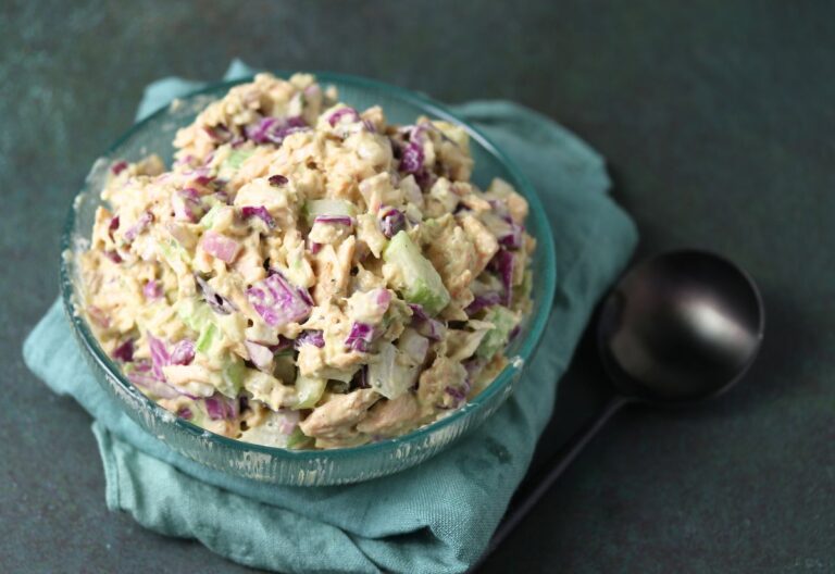 A glass bowl of chicken salad with chopped celery and red onion sits on a green cloth next to a black spoon.
