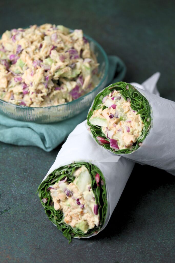 A spinach wrap filled with tuna salad, red onion, and cucumber, cut in half and wrapped in paper, with a bowl of tuna salad in the background.